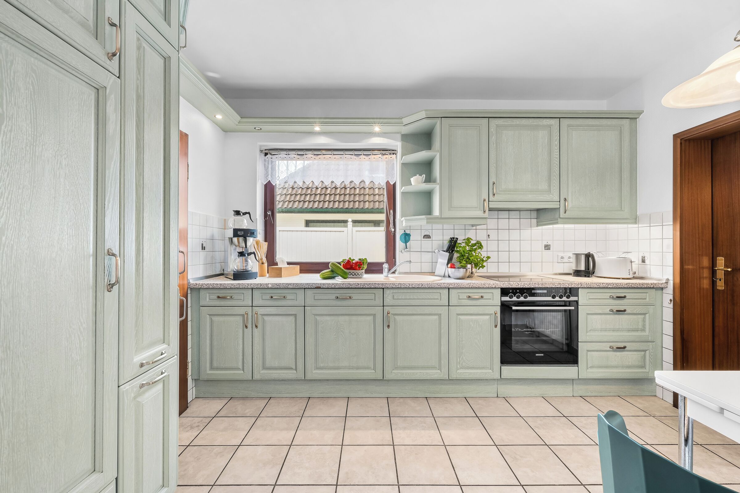 Eine Küche mit hellgrünen Schränken, einer gefliesten Spritzschutzwand, einem eingebauten Backofen und einem Fenster mit Vorhang.