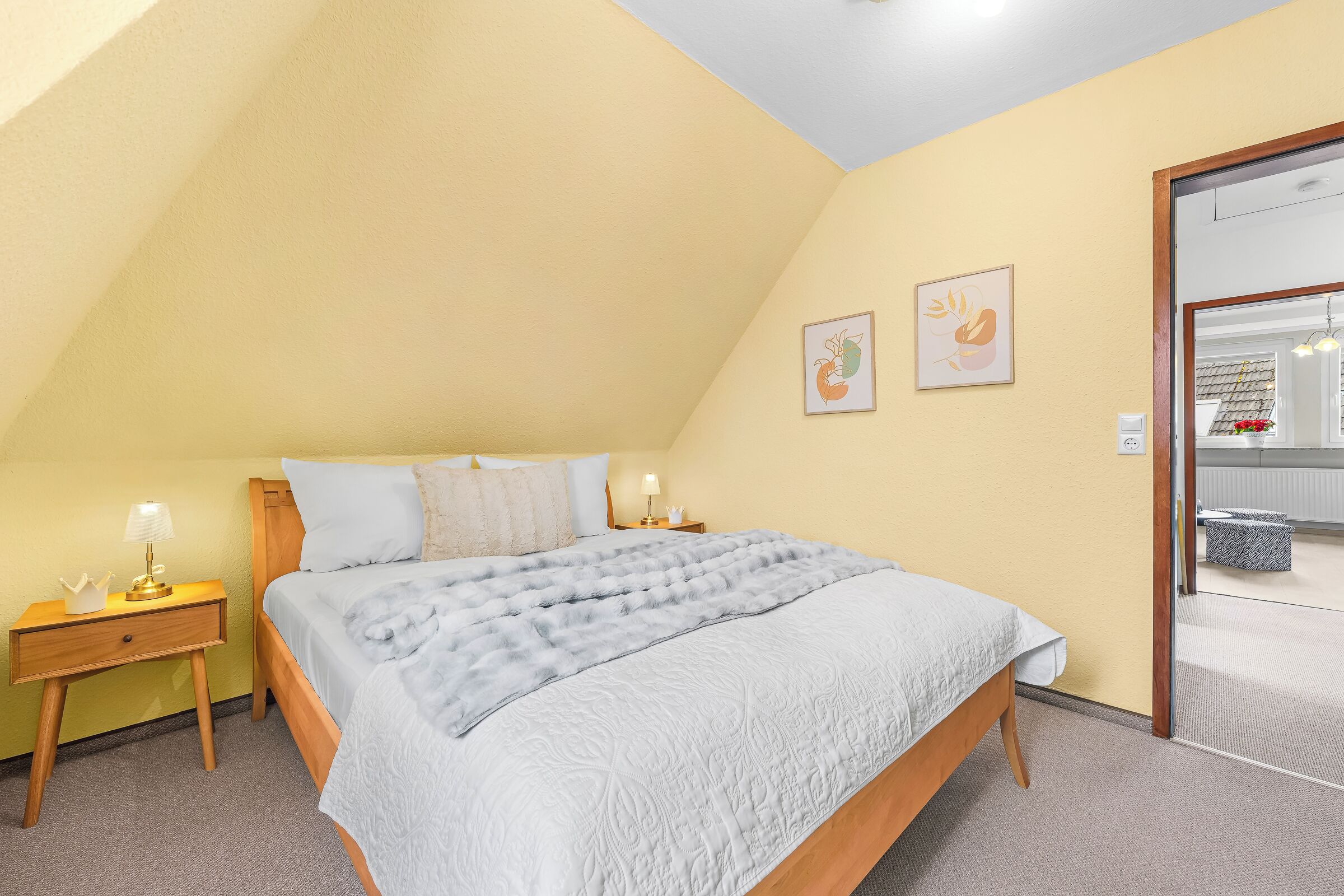 Ein Schlafzimmer mit geneigter Decke, einem Bett mit grauer Steudecke, zwei Nachttischen mit Lampen sowie zwei gerahmten Bildern an der Wand.
