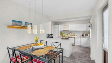 Een moderne keuken met witte kastjes, een eethoek met een glazen tafel, en een hangende lamp.