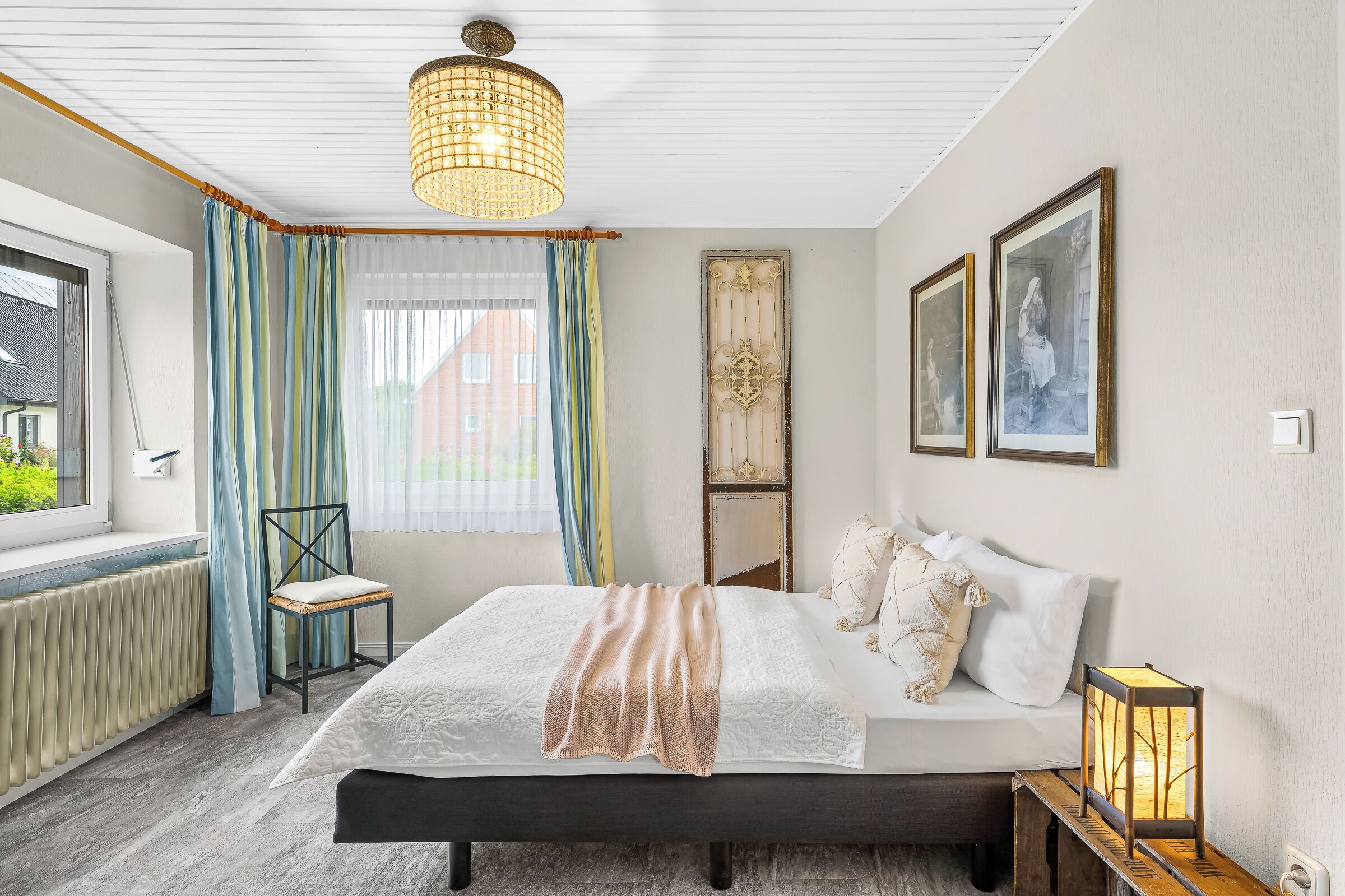 Een slaapkamer met een bed, een stoel, een raam met gordijnen, een radiator en een wandlamp.