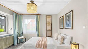 Ett sovrum med en säng, en stol, ett fönster med gardiner, ett element och en väggmonterad lampa.