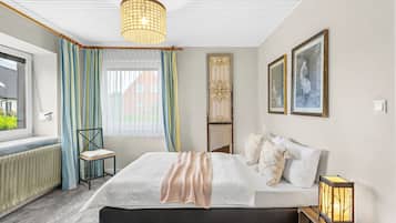 Een slaapkamer met een bed, een stoel, een raam met gordijnen, een radiator en een wandlamp.