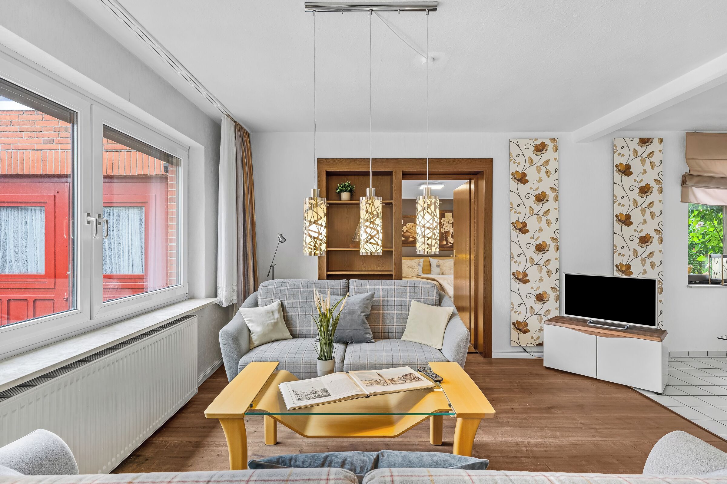 Een moderne woonkamer met een bank, een salontafel, een televisie en een raam met uitzicht op een rode gevel.