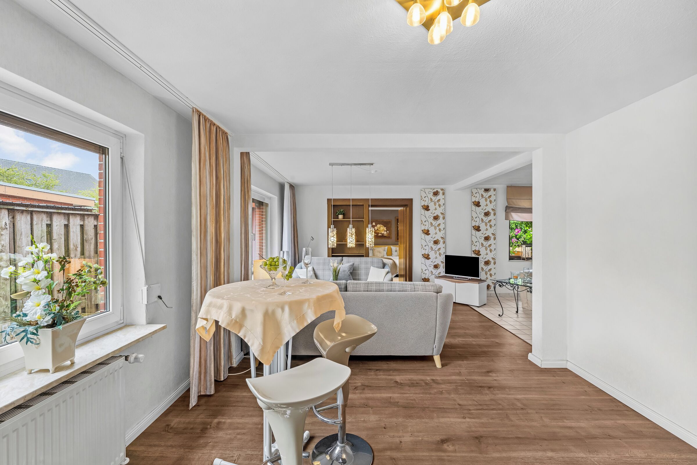 Ein modernes Wohnzimmer mit einer Couch, einem Esstisch mit Stühlen, einem Fernseher und einem großen Fenster mit Vorhängen.