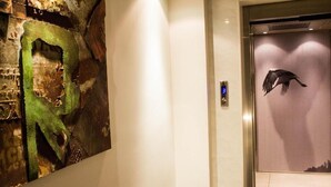 A hallway with a green abstract art piece on the left and an elevator with a bird silhouette on the right. - Hotel ILIANA (Filippiada)