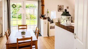A dining area with a wooden table and chairs, a window with curtains, and a view of greenery outside. - Lütt-Huus — for you & your four-legged friend (Blomberg)