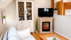 A living room with a sofa, a television, a cabinet with glass doors, and a fireplace. - Lütt-Huus — for you & your four-legged friend (Blomberg)