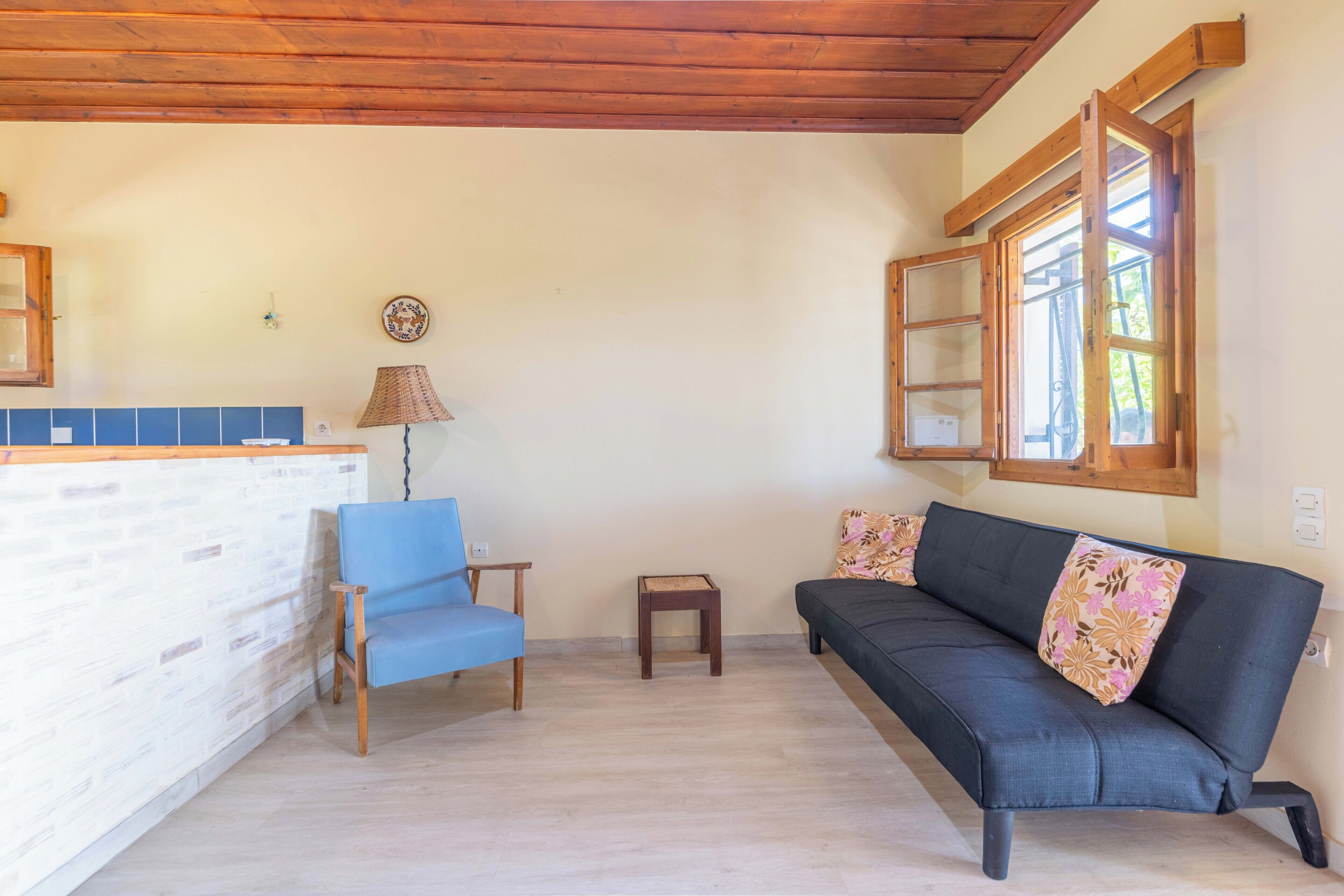 A living room with a blue chair, a sofa with floral cushions, a small table, and a window with wooden shutters.