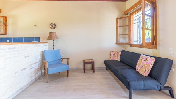 Ein Wohnzimmer mit blauem Sessel, einer Couch mit floralen Kissen, einem kleinen Tisch und einem Fenster mit Holzläden.