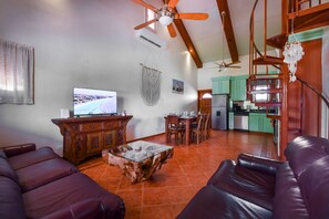 Ein Wohnzimmer mit Fernseher, Holzmöbeln, Deckenventilator und Küchenzeile mit grünen Schränken.