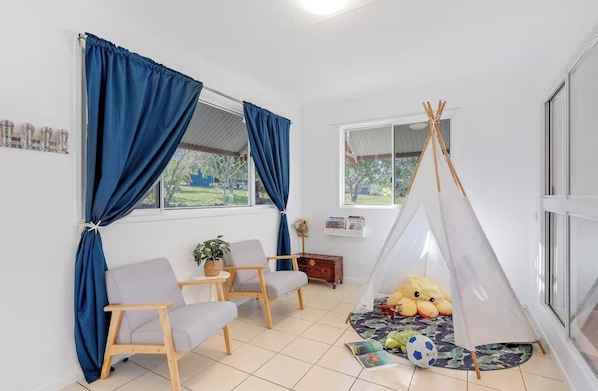 Una habitación con un ventanal grande, cortinas azules, una silla de madera, una mesita con una planta, un oso de peluche y un tipi.