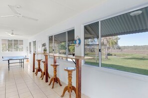 A room with a ping pong table, wooden stools, large windows, ceiling fans, and a view of a grassy area. - The big catch! - 3.5 acres - pets! - boats - fishing and crabbing! (Rodds Bay)