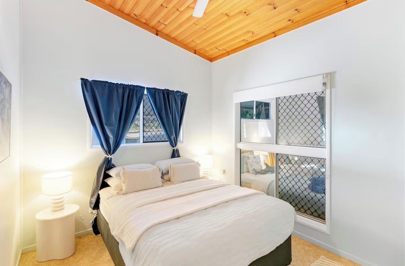Ein Schlafzimmer mit einem Bett, einem Nachttisch, einem Fenster mit Vorhängen und einer Decke mit Holzbalken.