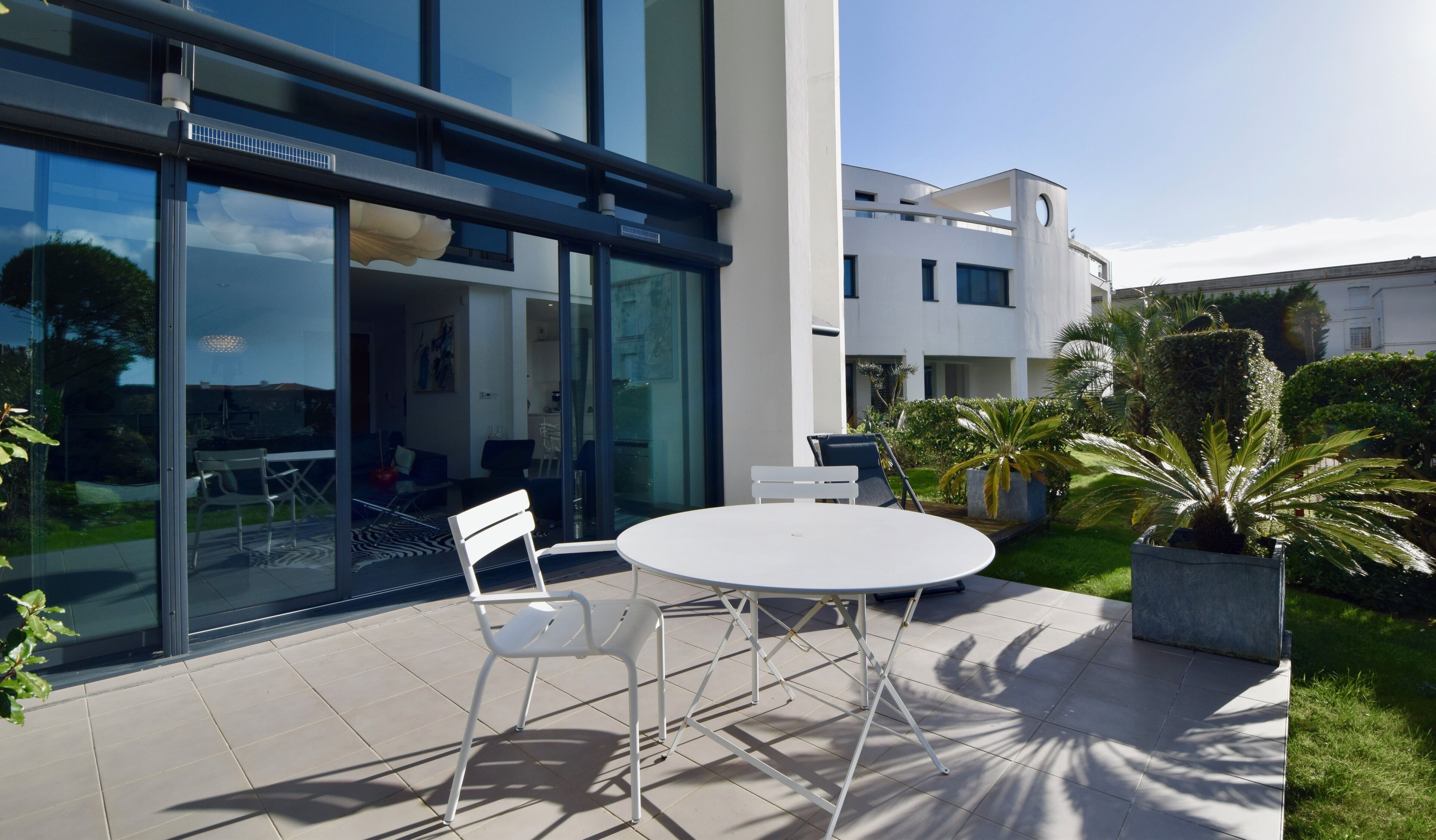 Een terras buiten met een ronde tafel en stoelen, een glazen deur en een modern gebouw op de achtergrond.