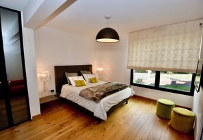A modern bedroom with a large bed, a ceiling light, a window with blinds, and a view of the outdoors. - PRESTIGE RANGE - VACATION RENTALS IN ROYAN AND PONTAILLAC - 2-BEDROOM APARTMENT (Royan)