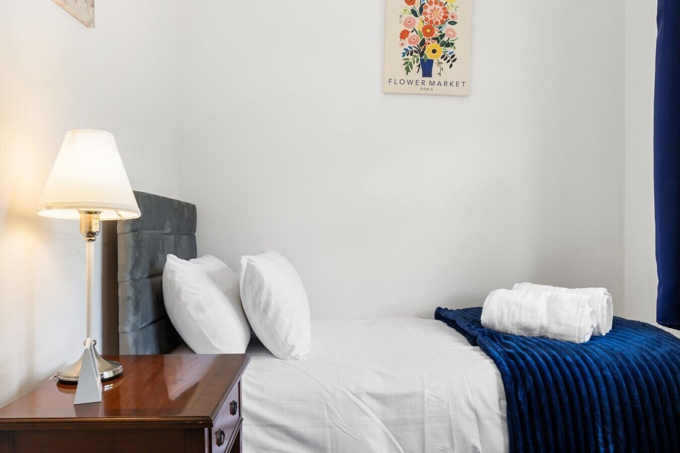 A hotel room with a bed, a bedside table with a lamp, a blue blanket, and a wall with a floral artwork.