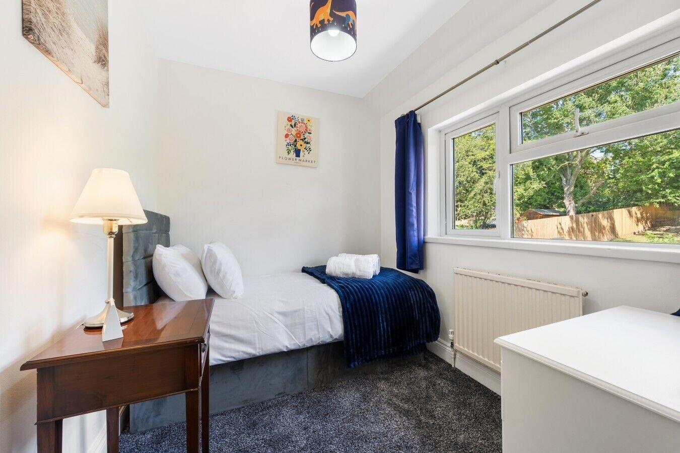 A hotel room with a bed, a desk, a lamp, a radiator, and a window with a view of trees.