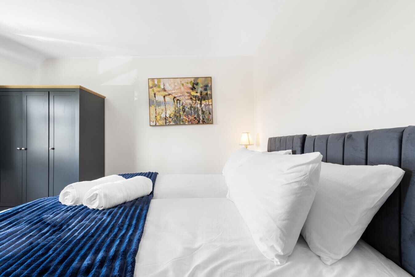 A hotel room with a bed, a painting, a wardrobe, and a bedside lamp.