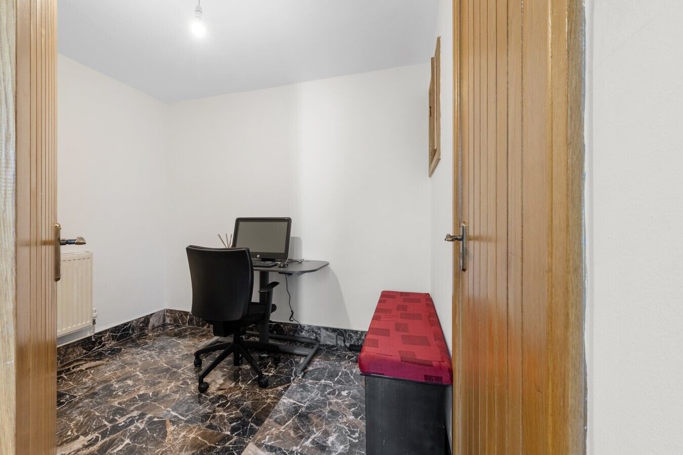 A room with a desk, chair, and a computer. There is a radiator and a door with a wooden frame.