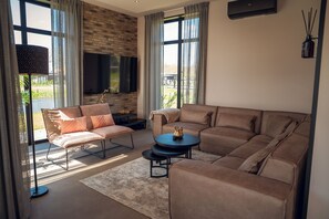 Ein modernes Wohnzimmer mit einem Ecksofa, einer Liege, einem Flachbildfernseher an einer Ziegelwand und großen Fenstern mit Vorhängen.