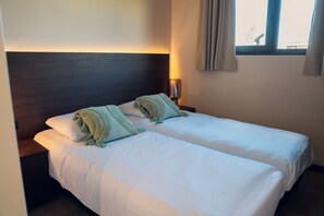 Ein Doppelbett mit zwei Kissen und einer Decke, ein Nachttisch mit Lampe sowie ein Fenster mit Vorhängen.