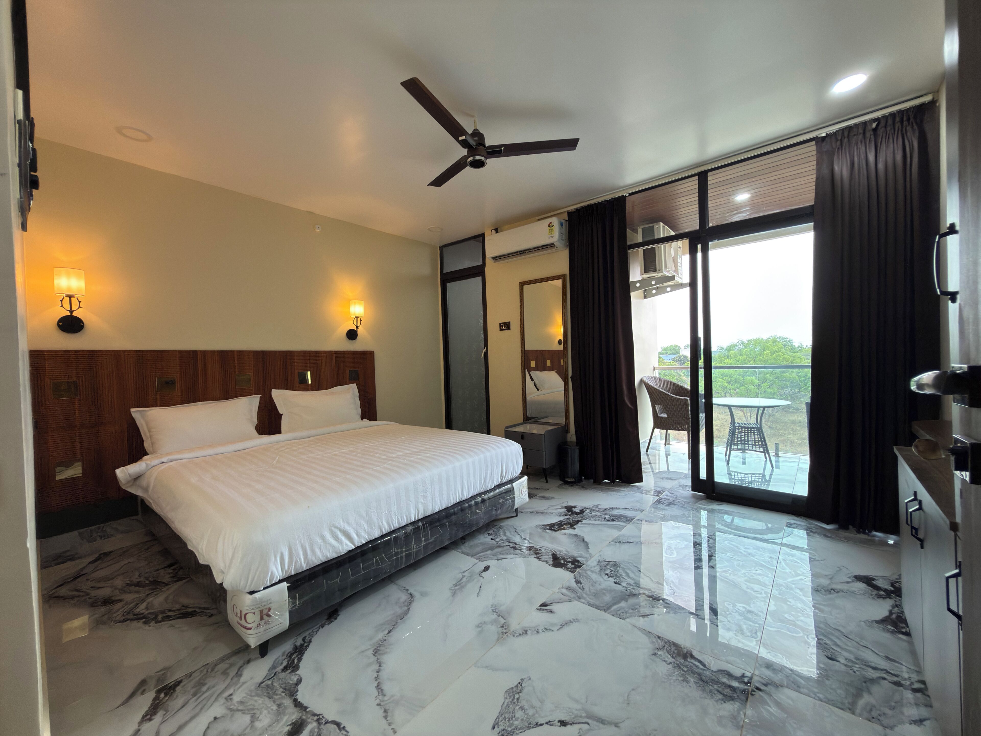 A hotel room with a bed, ceiling fan, marble flooring, and a balcony with a table and chairs.