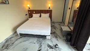 Quarto de hotel com cama, duas luminárias na parede, ventoinha no teto, piso com padrão marmorizado, espelho e ar-condicionado.