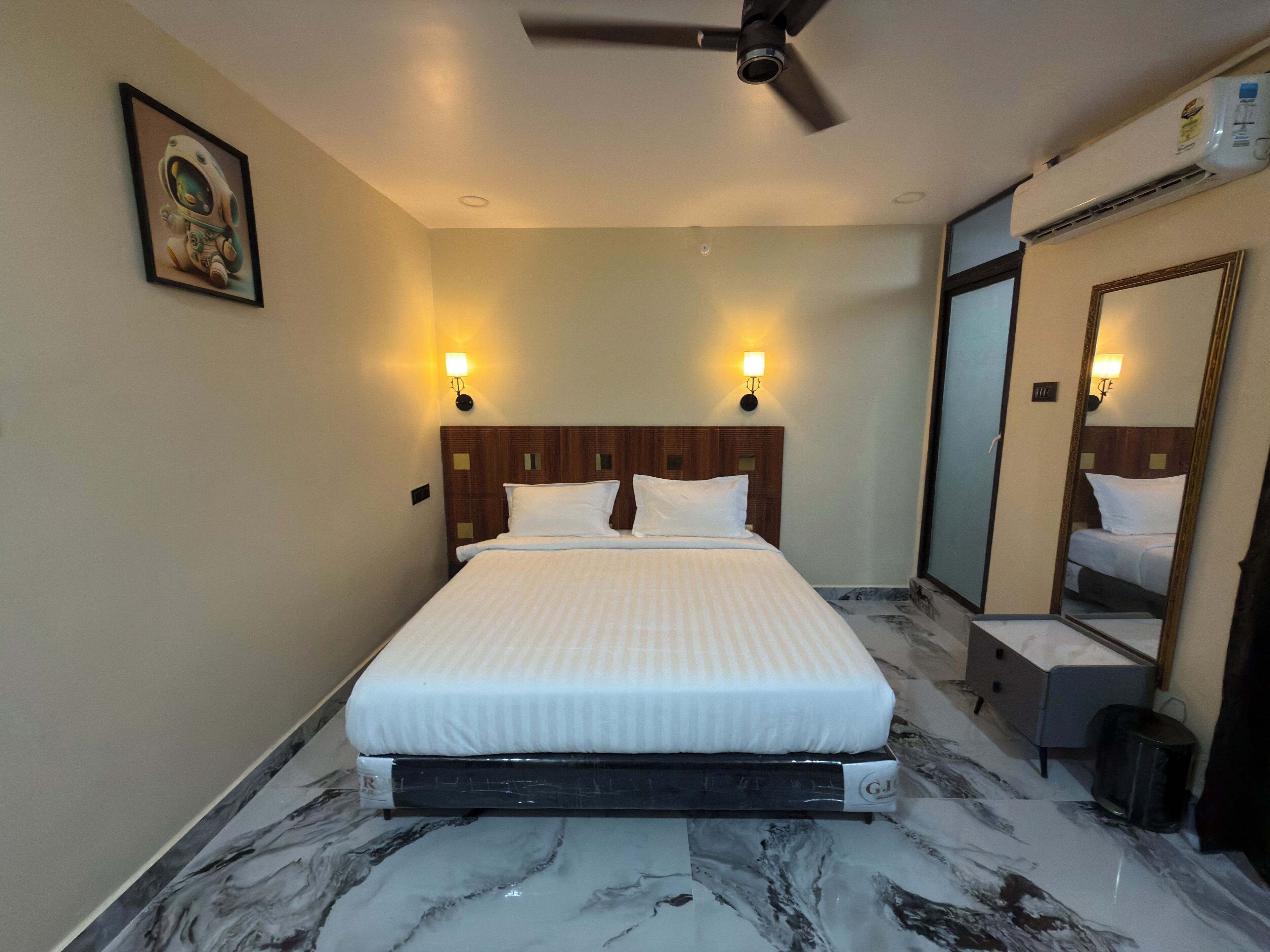 A hotel room with a bed, a ceiling fan, a wall-mounted air conditioner, a framed picture, and a large mirror.