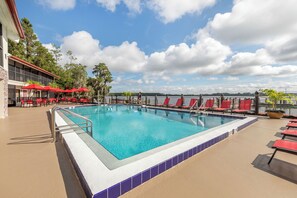 A swimming pool with a tiled edge, surrounded by red chairs and umbrellas, with a view of the sky and trees in the background. - Orlando Family Getaway | Resort Condo Minutes from Disney (Orlando)