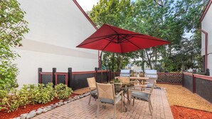 Een terras buiten met een rode parasol, een metselwerk vloer en een barbecue.