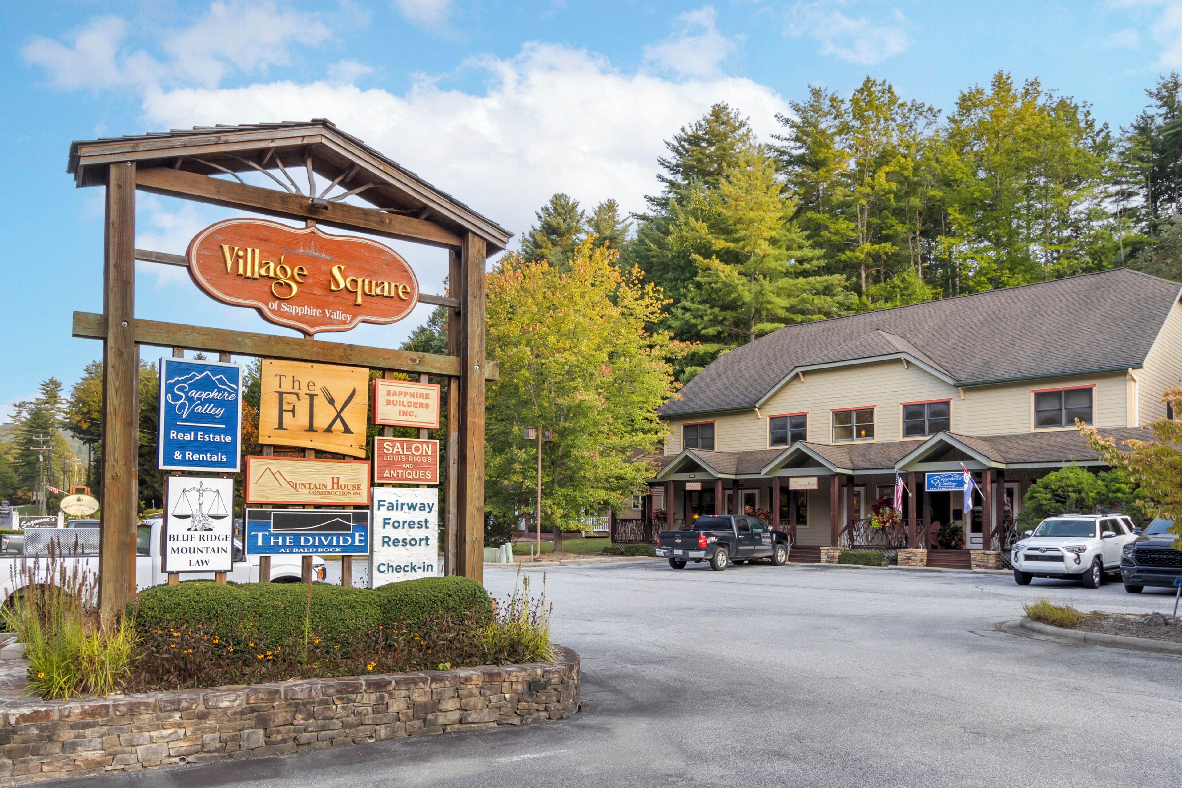 Une enseigne indiquant l’entrée de « Village Square of Sapphire Valley », avec divers panneaux indiquant les commerces, un bâtiment doté d’un porche et des voitures garées.