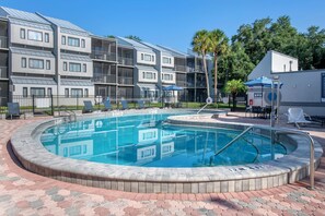 A swimming pool with a surrounding brick walkway, a covered seating area, and a multi-story building with balconies in the background. - Disney Next Door, Family Time Everywhere | 2BR Resort Villa (Kissimmee)
