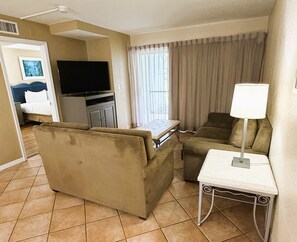 A hotel room with a sofa, a small table with a lamp, a television, and a window with curtains. - Disney Next Door, Family Time Everywhere | 2BR Resort Villa (Kissimmee)