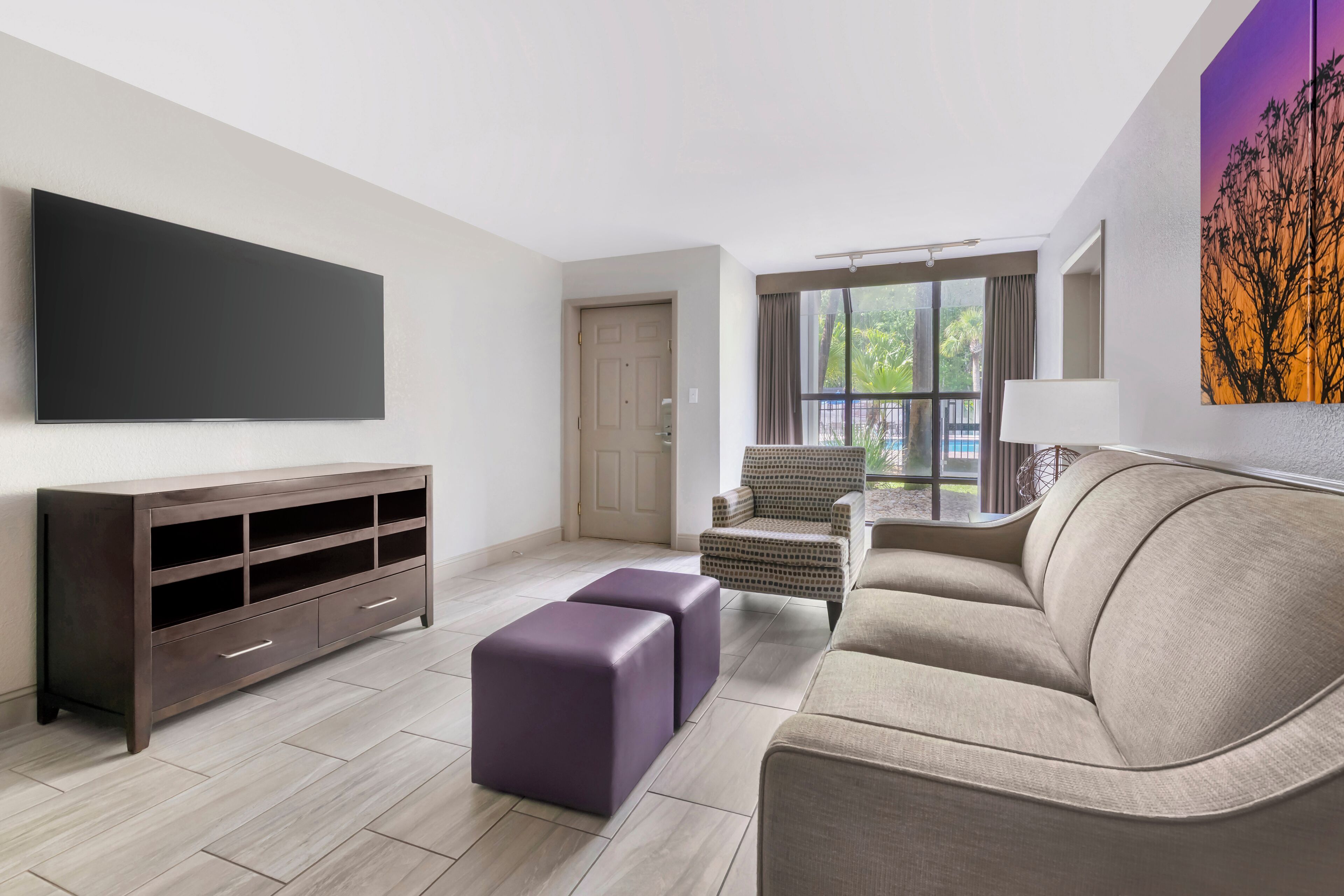 A modern living room with a flat-screen TV, a dark wooden entertainment unit, a beige sofa, and a purple ottoman.