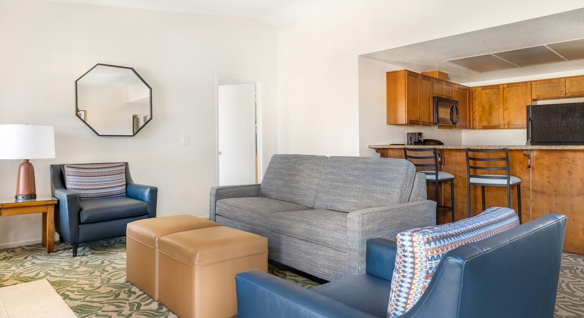 A compact living space with a sofa, ottoman, and chairs, a kitchenette with wooden cabinets, and a large mirror on the wall.