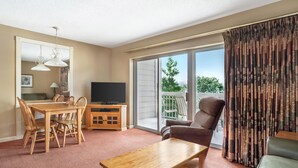 Ein Wohnzimmer mit Holztisch, Stühlen, einer Couch, einem Fernseher und einer Glastür zum Balkon.
