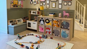 Ein Kinderzimmer mit einer Modelleisenbahn auf einem gemusterten Teppich, Regalen mit Büchern und Spielzeug sowie gerahmten Bildern an der Wand.