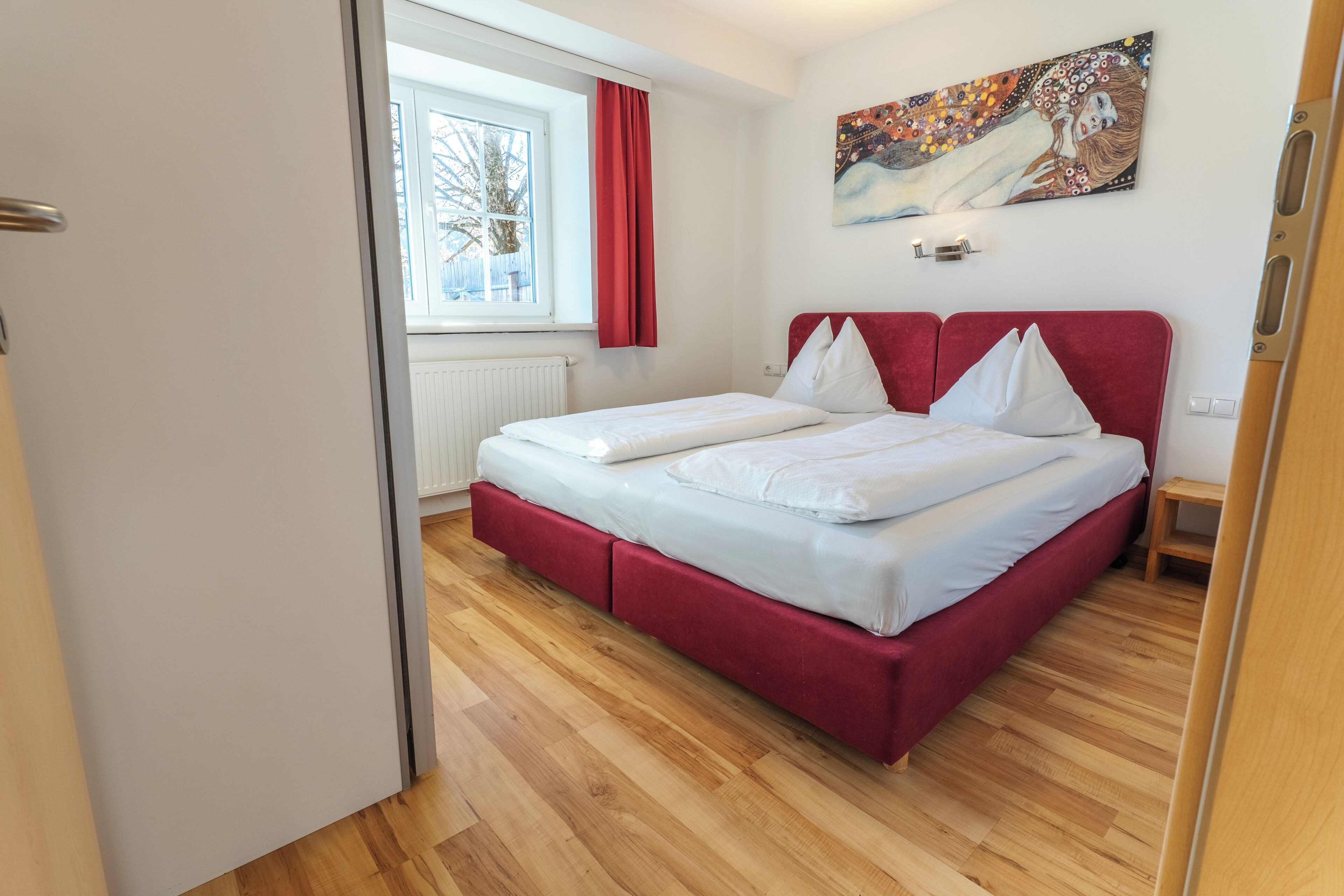 Un dormitorio con cama, ventana con cortinas, radiador y un cuadro en la pared.