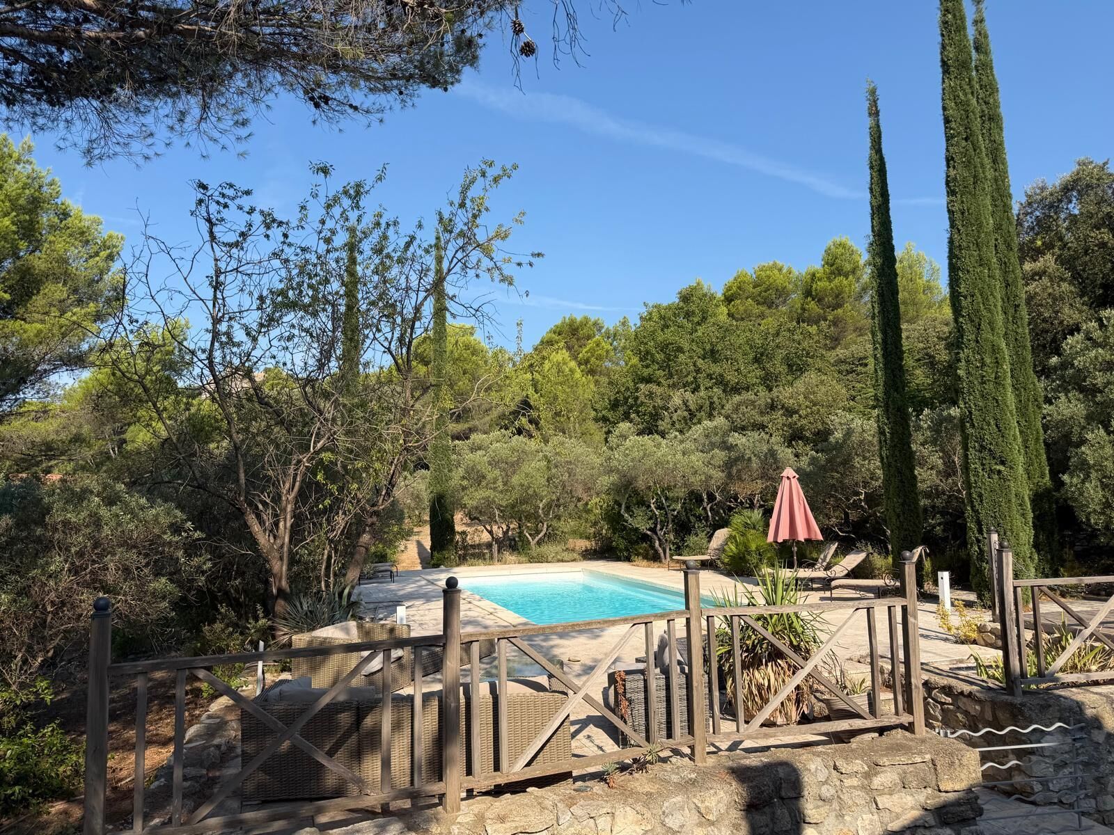 A pool surrounded by a wooden fence, with a red umbrella and lounge chairs nearby.