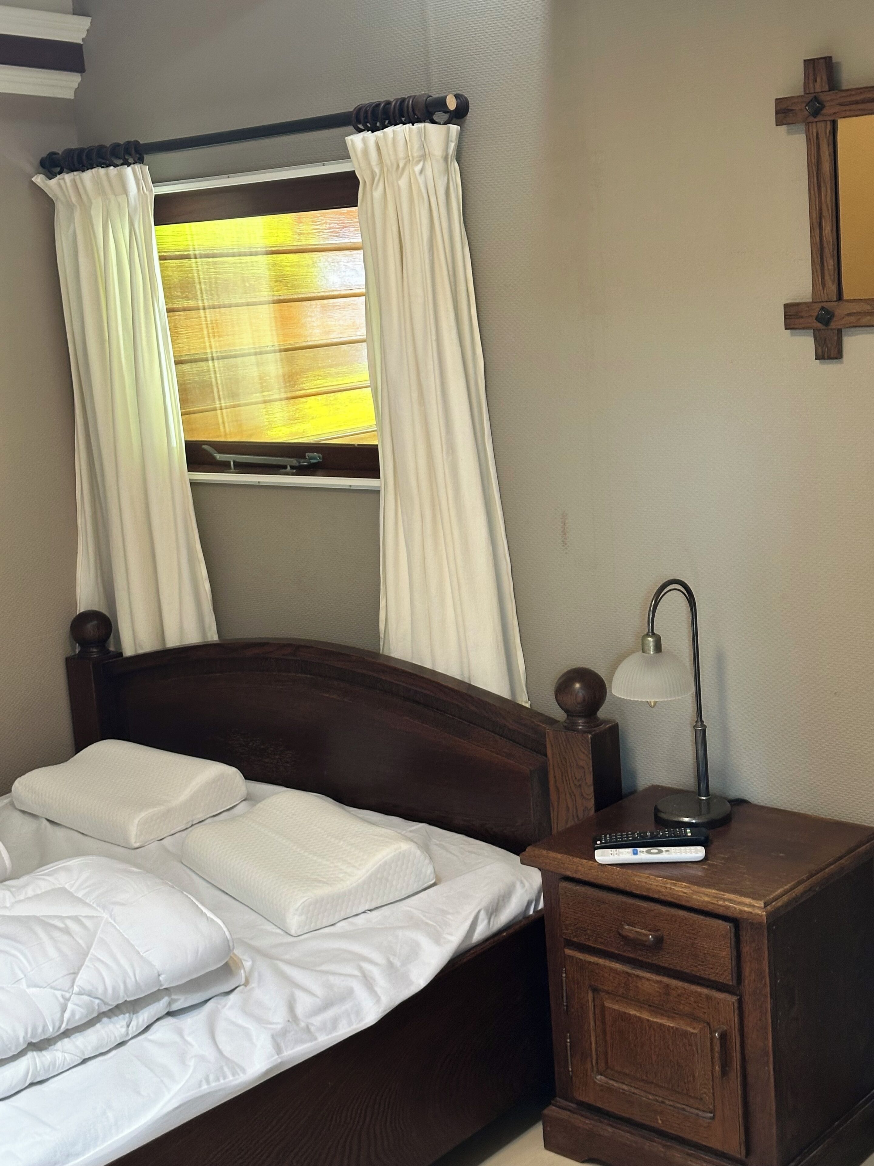 Un lit avec une literie blanche, une tête de lit en bois, une table de chevet avec une lampe et une fenêtre avec des rideaux.