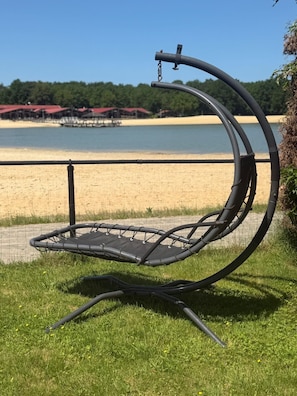 A curved outdoor seating area with a metal frame and a black seat cushion, located on grass with a view of a beach and water in the background.