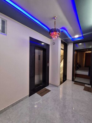 Un pasillo moderno con piso de mármol, un elevador negro, una lámpara decorativa y una puerta que da a una habitación.