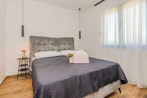 Een slaapkamer met een groot bed, een nachtkastje, een vaas met bloemen en een raam met dunne gordijnen.