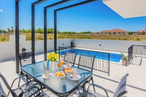 A glass-top table with chairs, a vase with yellow flowers, a glass of orange juice, and a plate of food on a balcony overlooking a swimming pool. - Villa Caelora-Private Villa With Pool-Pet Friendly (Privlaka)