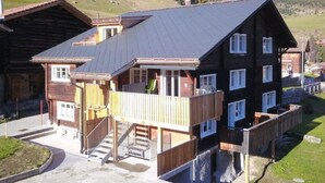 En tvåvånings chalet i trä med en balkong, en trappa och utsikt över ett bergigt landskap.