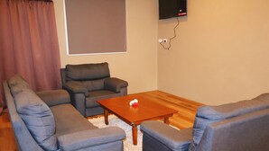 A living room with a sectional sofa, a single armchair, a wooden coffee table, and a flat-screen TV mounted on the wall. - Bula Accommodation (Nadi)