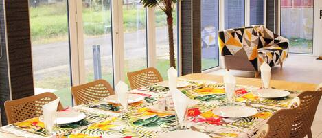 A dining area with a table set for four, surrounded by wicker chairs, and large windows offering a view of greenery and a road.