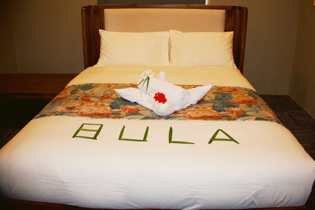 Un letto d'hotel con copriletto floreale, un asciugamano arrotolato con un fiore e la scritta 'BULA' sul copriletto.