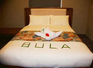 Un lit d’hôtel avec une couvre-lit à motifs floraux, une serviette roulée ornée d’une fleur, et le mot « BULA » inscrit sur la couvre-lit.