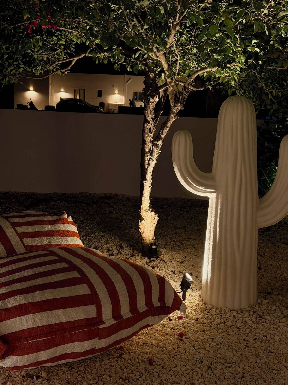 A cactus sculpture, a striped pillow, and a tree with a lit base in a nighttime outdoor setting.